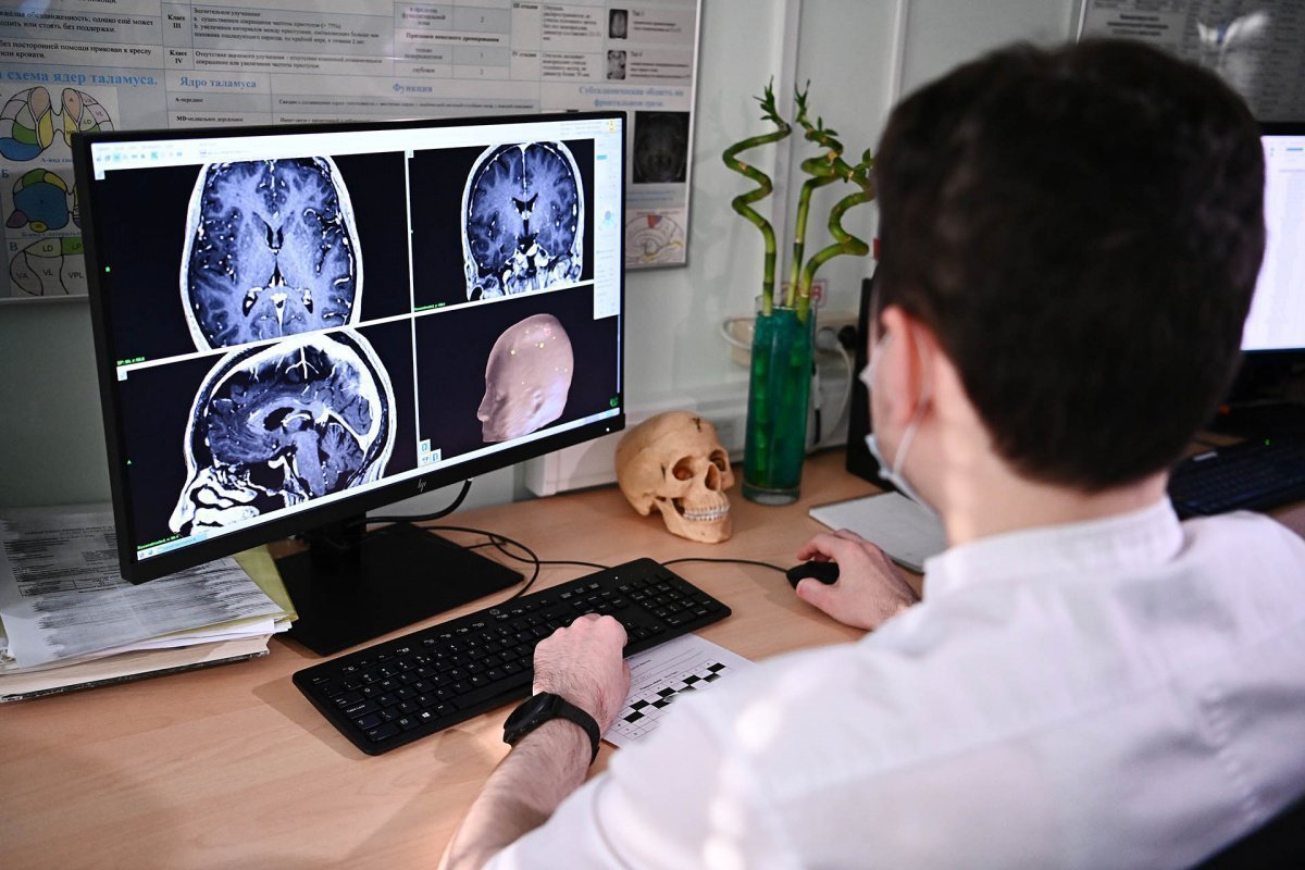 Медицинский работник за компьютером наблюдает за показаниями пациента помещенного в мрт на экране мозг в разрезе